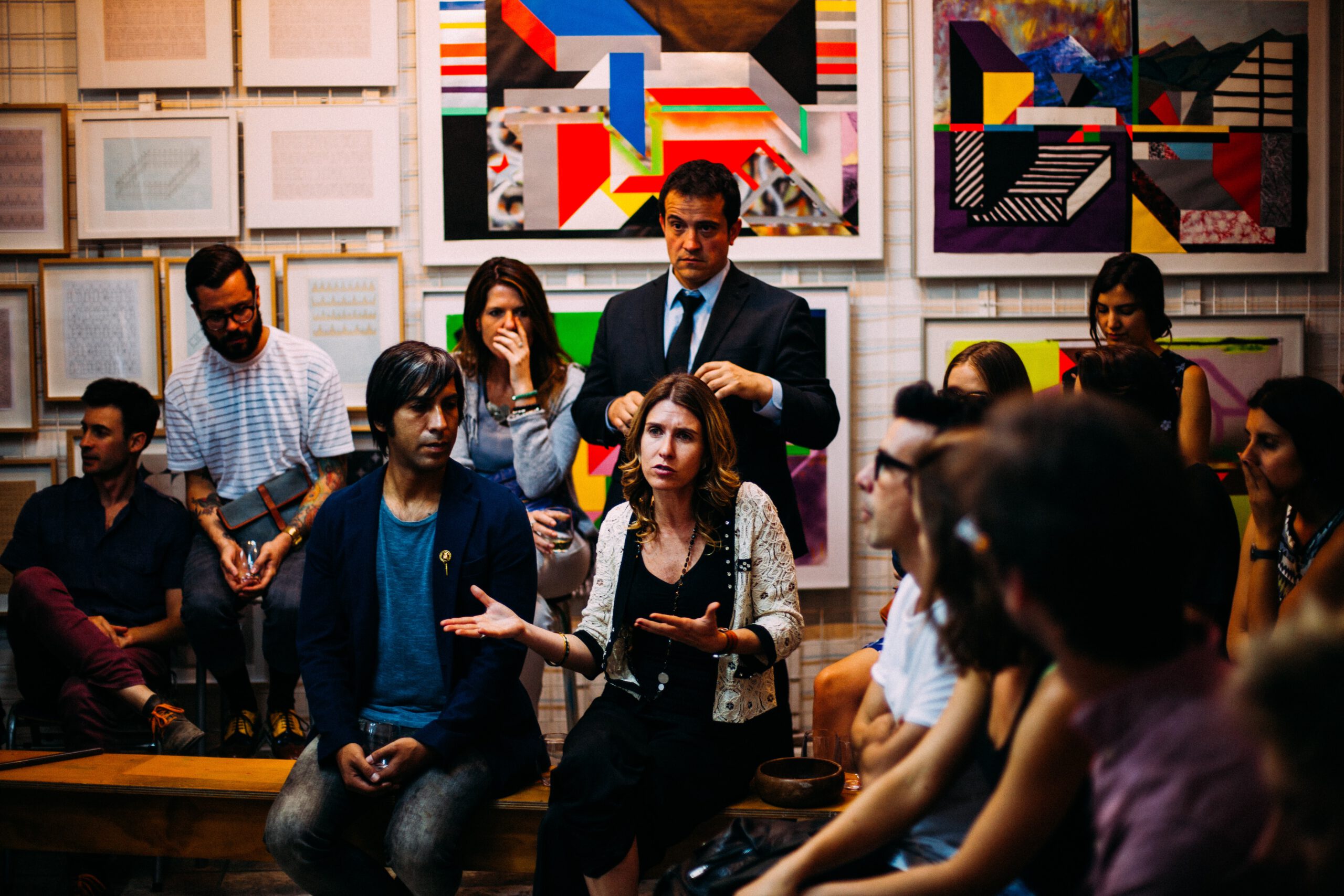 Foto: Eine Gruppe von 10 Menschen sitzen vor einer Wand mit bunten Kunstwerken und diskutieren. Eine Frau in der Mitte sagt etwas und hat die Arme vor sich fragend geöffnet.